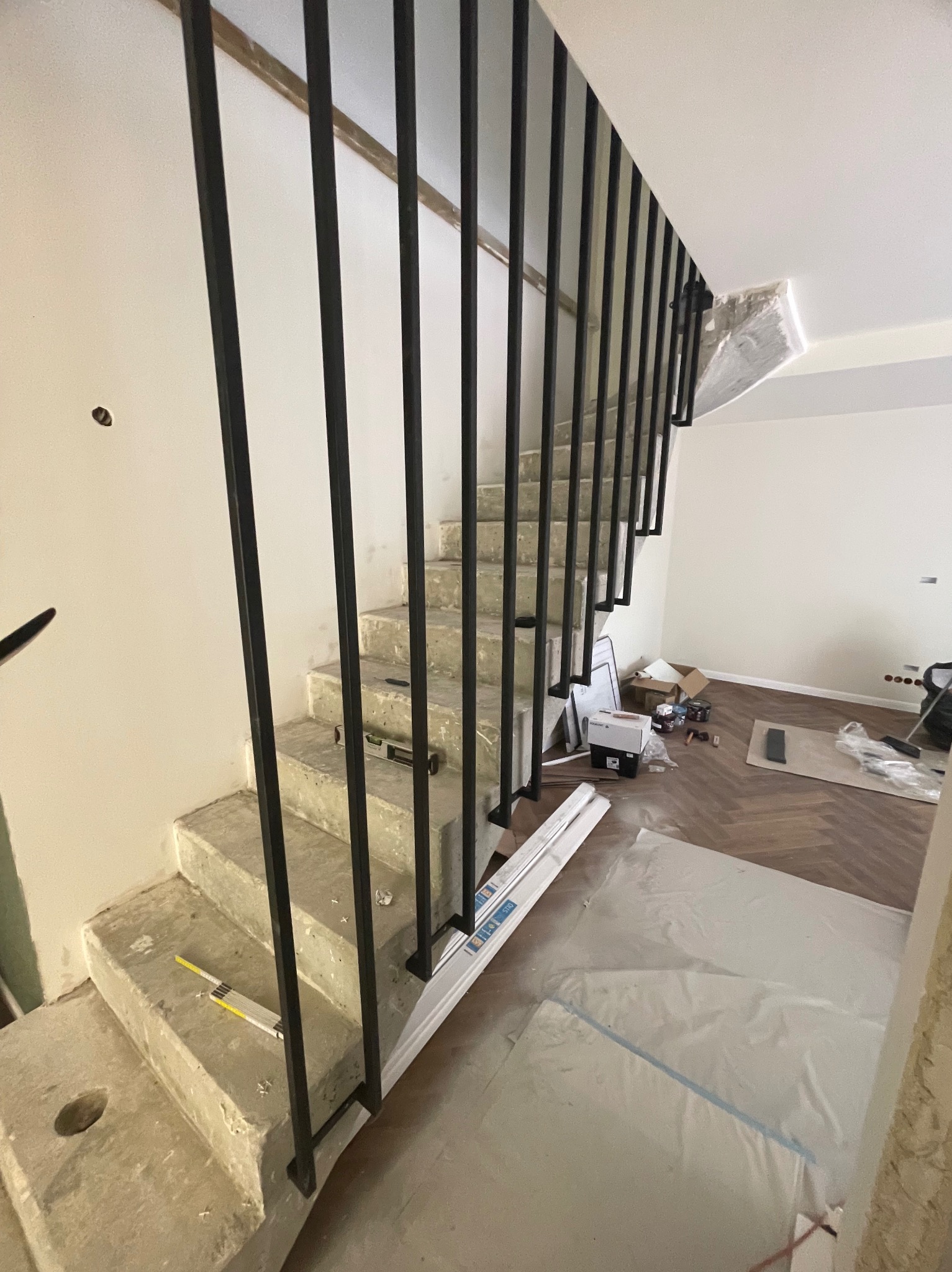 Betonowe schody wewnętrzne z czarną, minimalistyczną poręczą w trakcie montażu; widoczne narzędzia pomiarowe i zabezpieczenia podłogi.