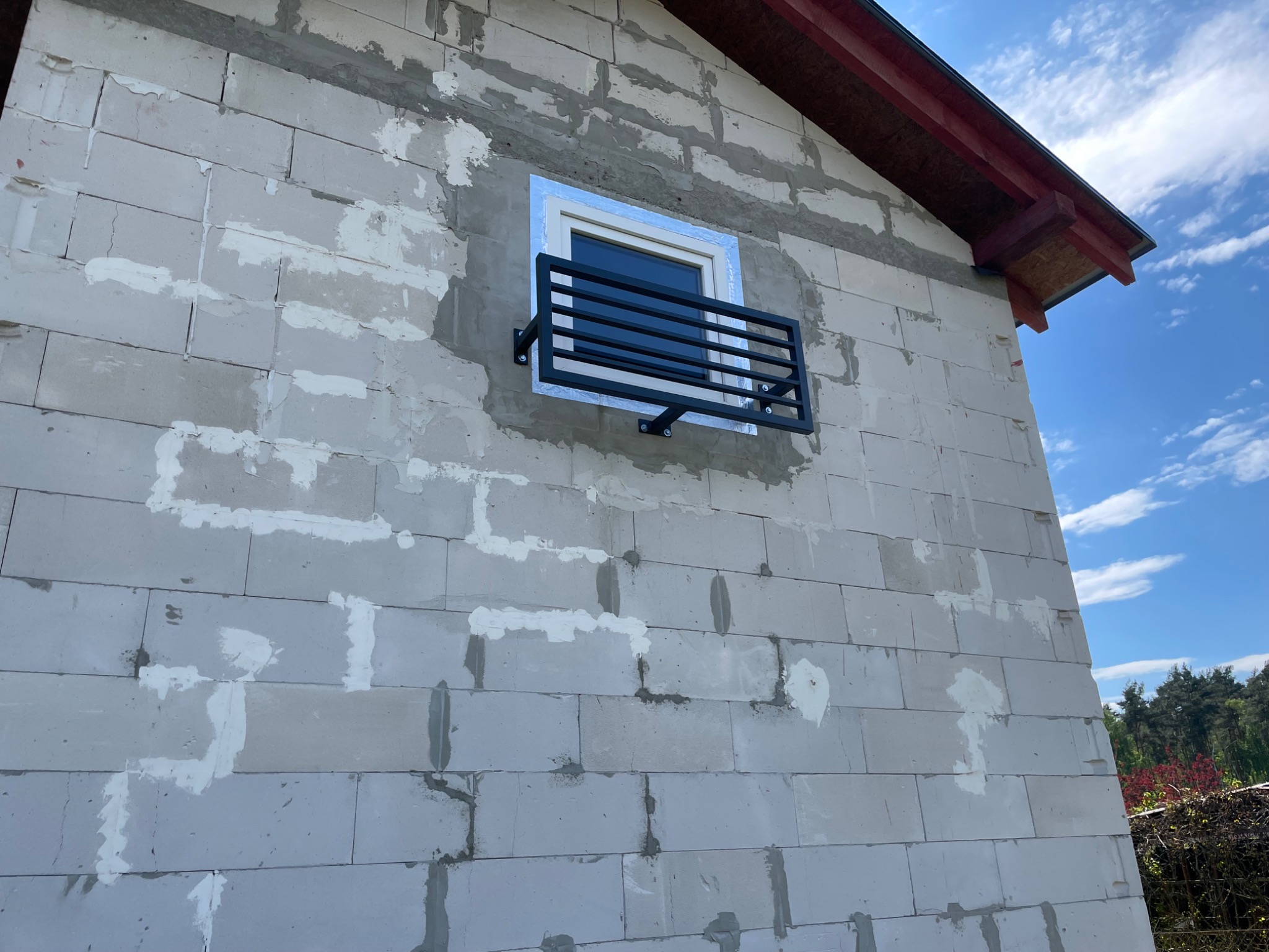 Czarna, metalowa barierka okienna zamontowana na surowej elewacji budynku z betonu komórkowego, zabezpieczająca okno na parterze.