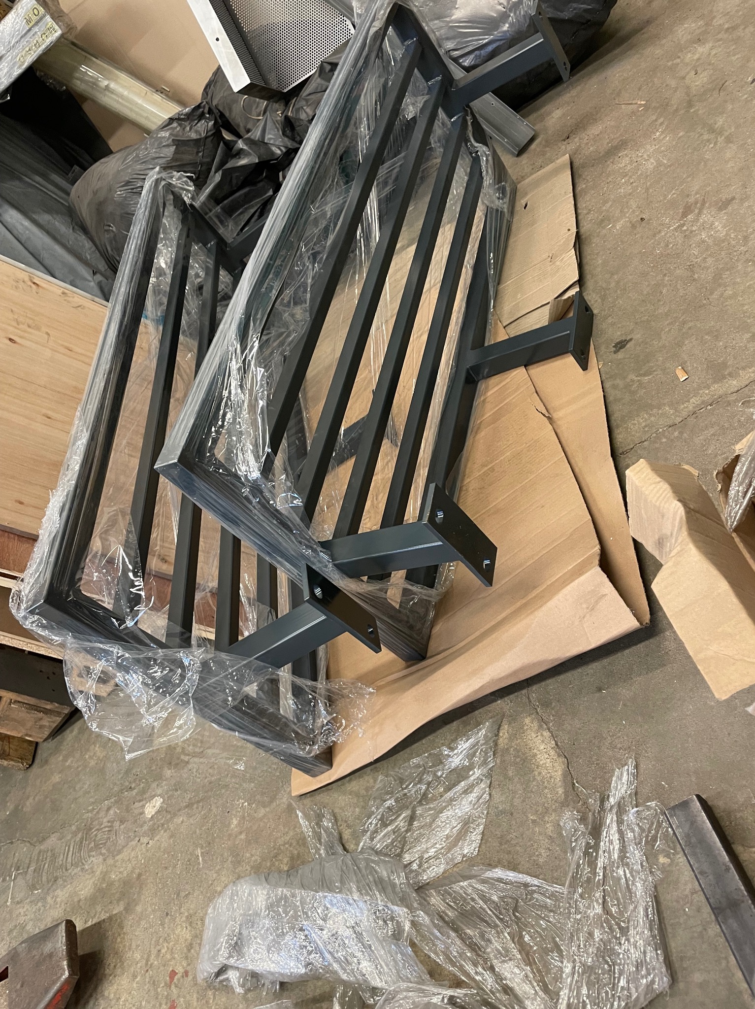 Elementy metalowe konstrukcji, prawdopodobnie balustrady lub schodów, zapakowane w folię ochronną i leżące na tekturze w warsztacie.