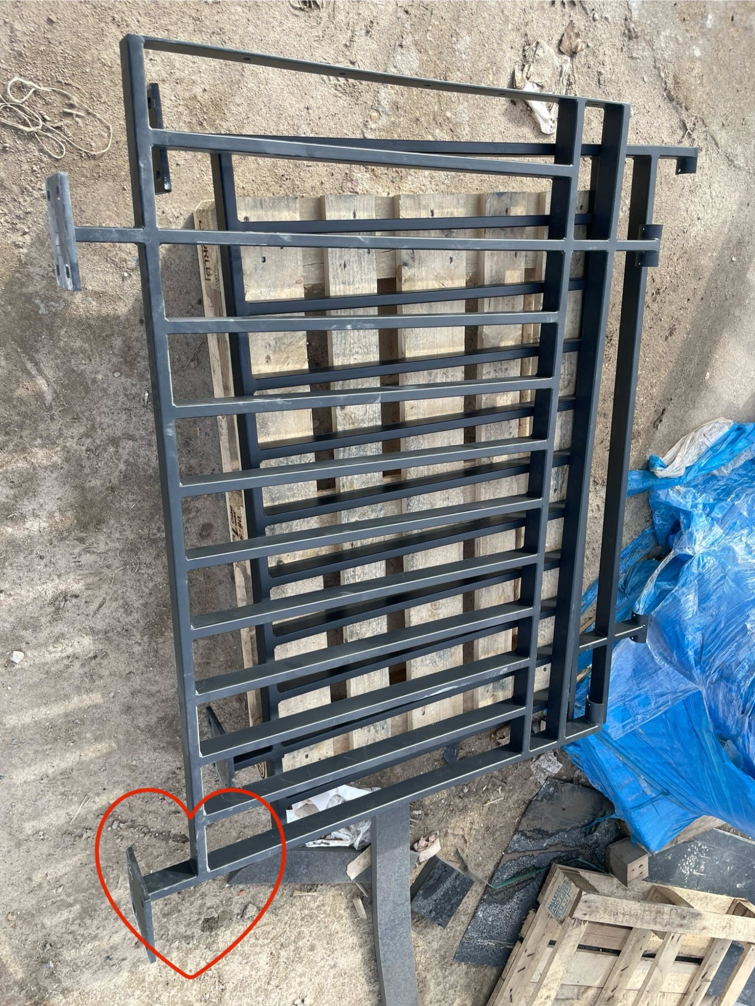 Surowa, spawana konstrukcja metalowa z płaskowników i profili kwadratowych w kolorze grafitowym, leżąca na betonowym podłożu i drewnianej palecie, prawdopodobnie element bramy lub ogrodzenia...