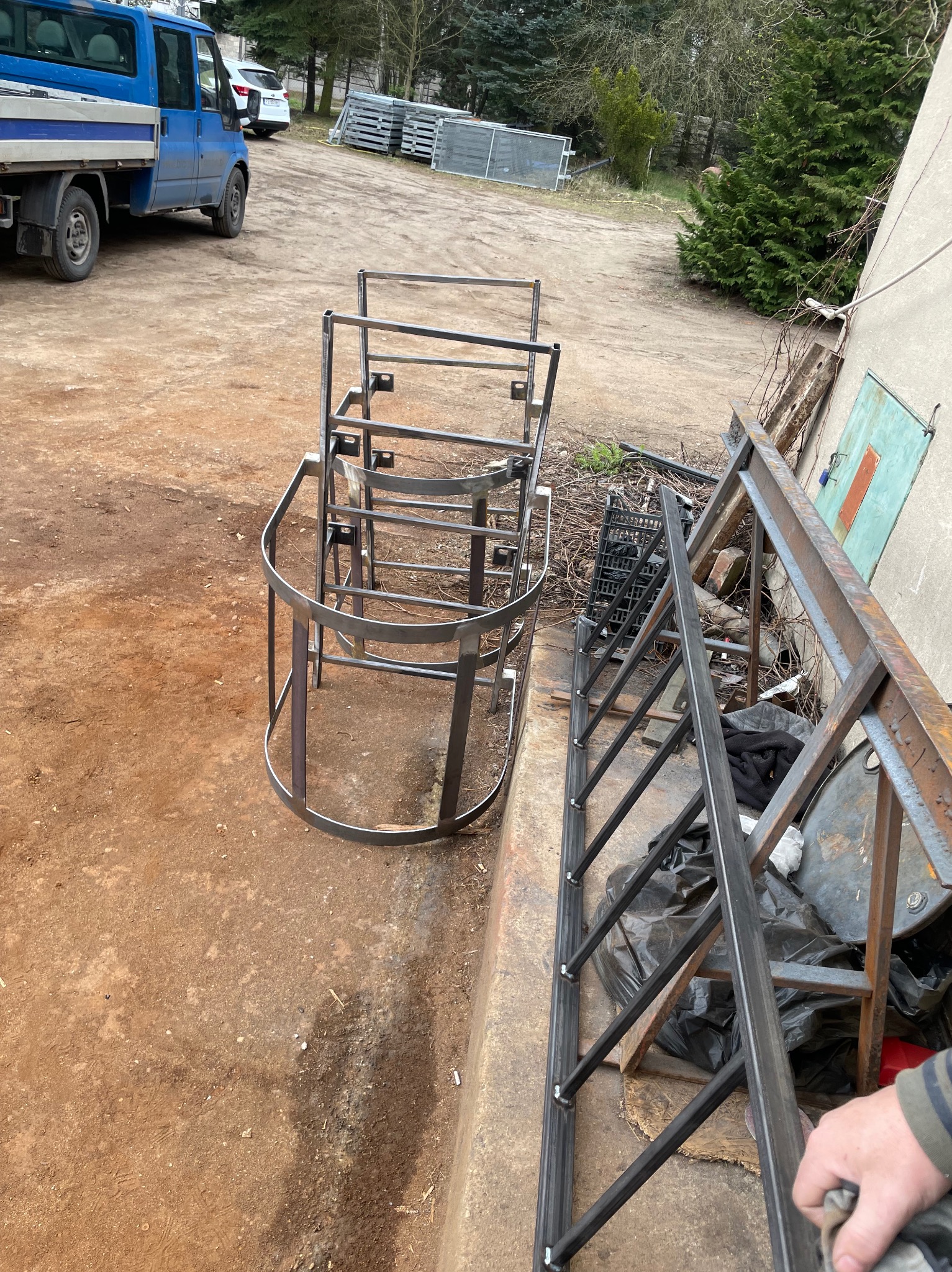 Surowe, metalowe konstrukcje schodowe i poręcze na placu budowy, widoczny niebieski samochód dostawczy i inne elementy metalowe w tle.