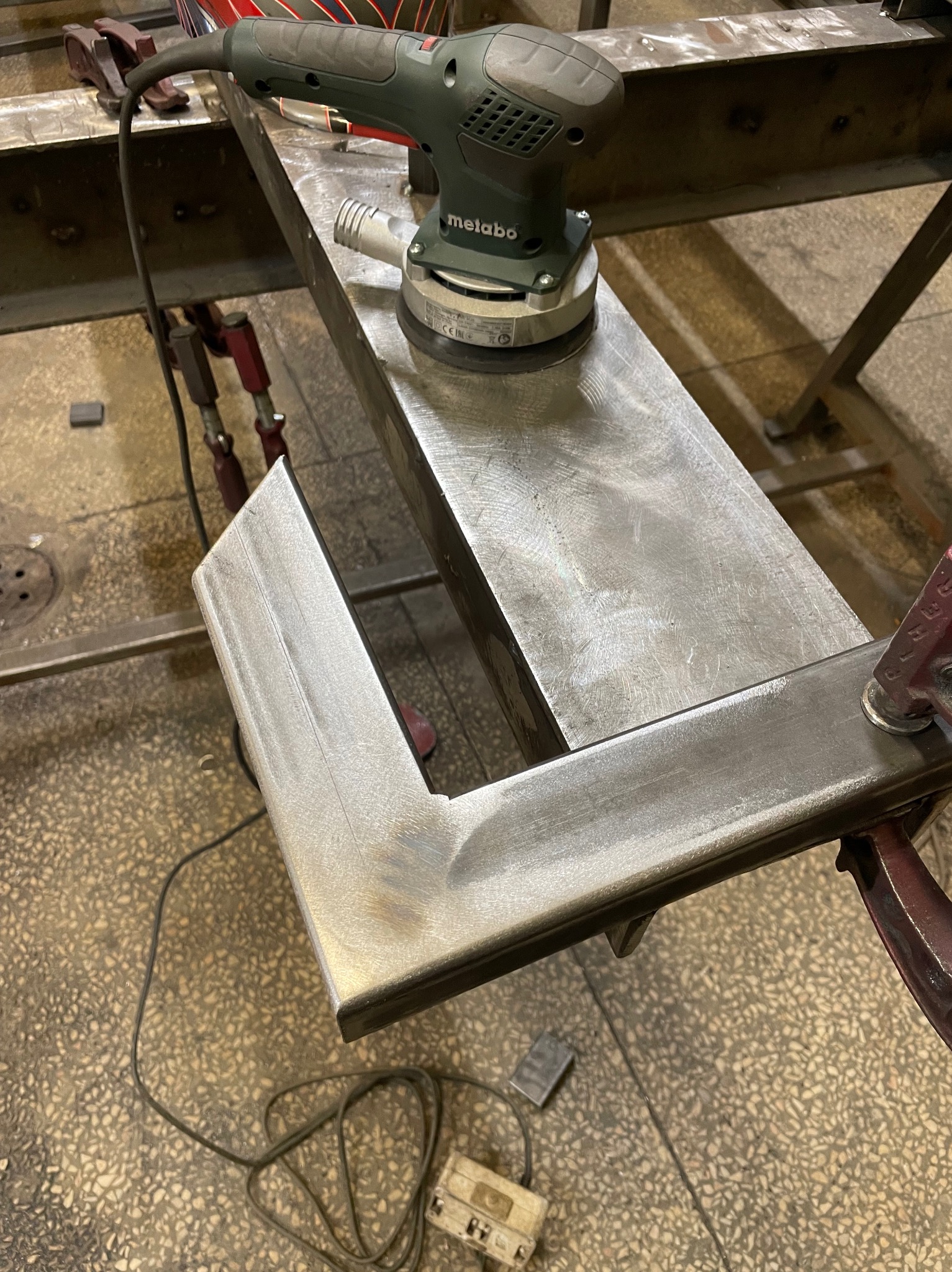 Szlifowanie metalowego profilu w kształcie litery U szlifierką oscylacyjną marki Metabo, zamocowanego klamrami do metalowej konstrukcji stołu warsztatowego.