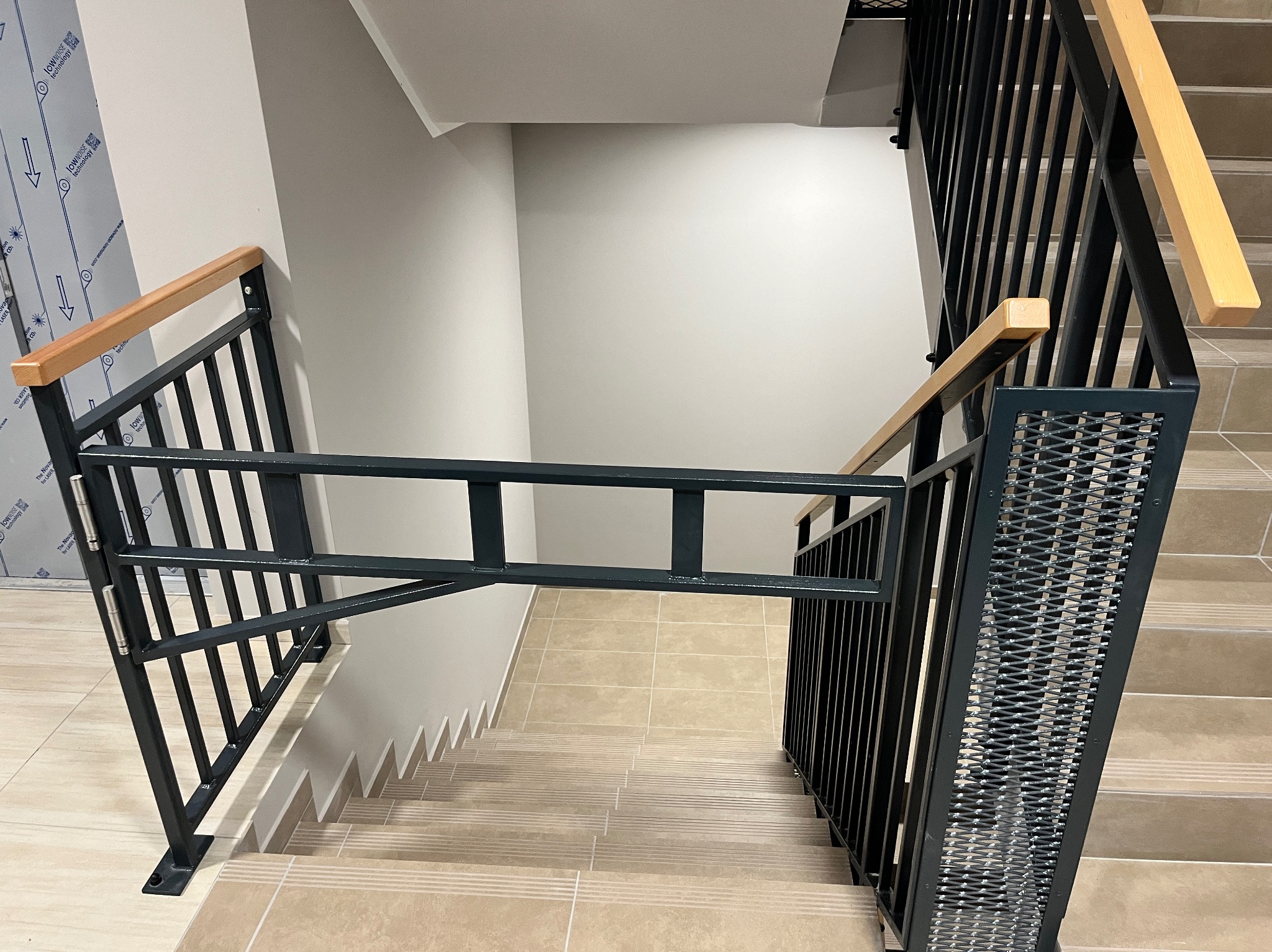 Klatka schodowa z metalowymi schodami i zabezpieczeniem w postaci barierki z drewnianymi poręczami, widok z góry na beżowe płytki schodowe i proste, nowoczesne wykończenie.