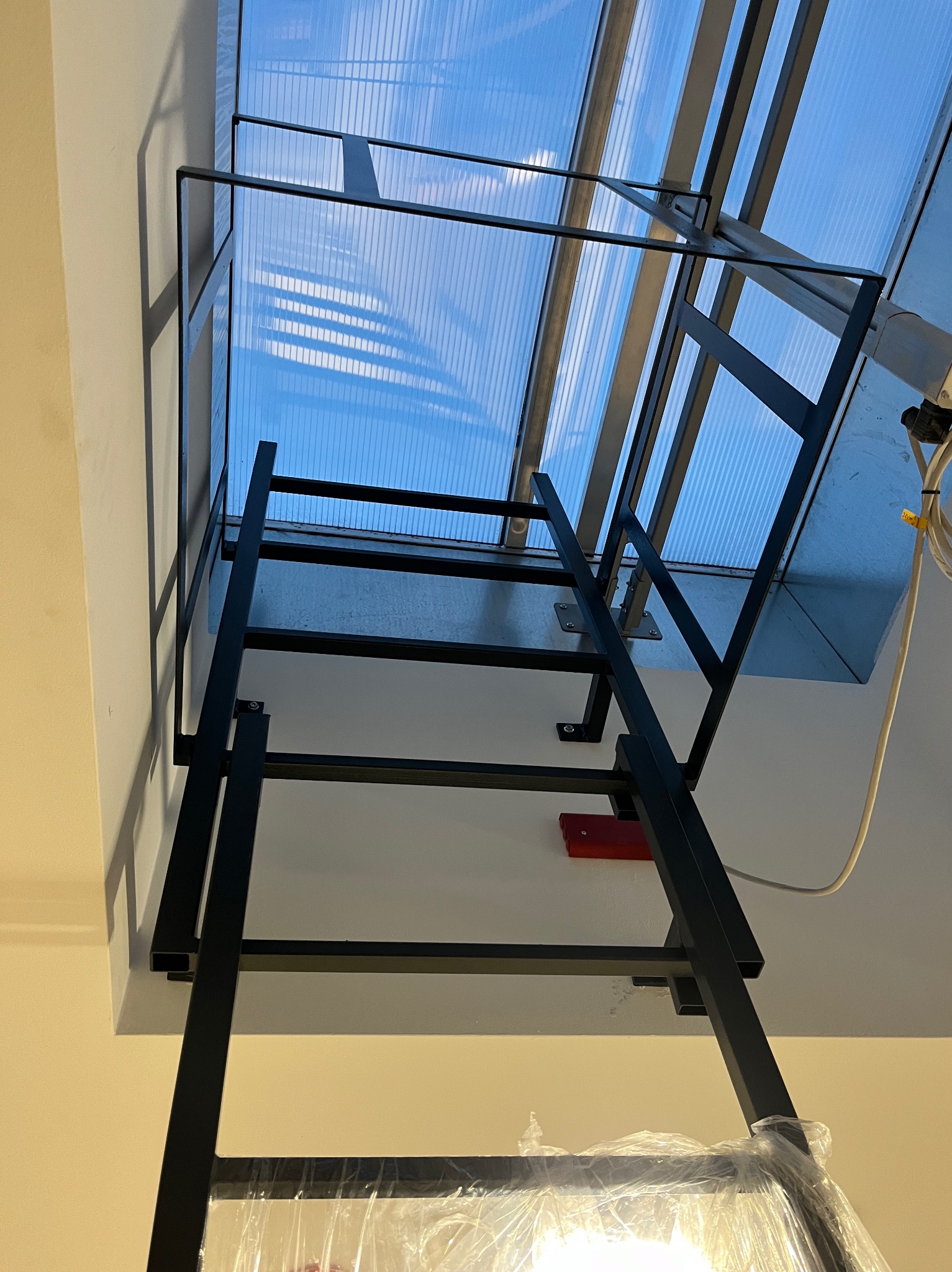 Czarna, metalowa drabina przymocowana do ściany i sufitu, prowadząca do świetlika z poliwęglanu. Widoczne mocowania drabiny do ściany i sufitu, oraz okablowanie elektryczne w pobliżu świetlika.