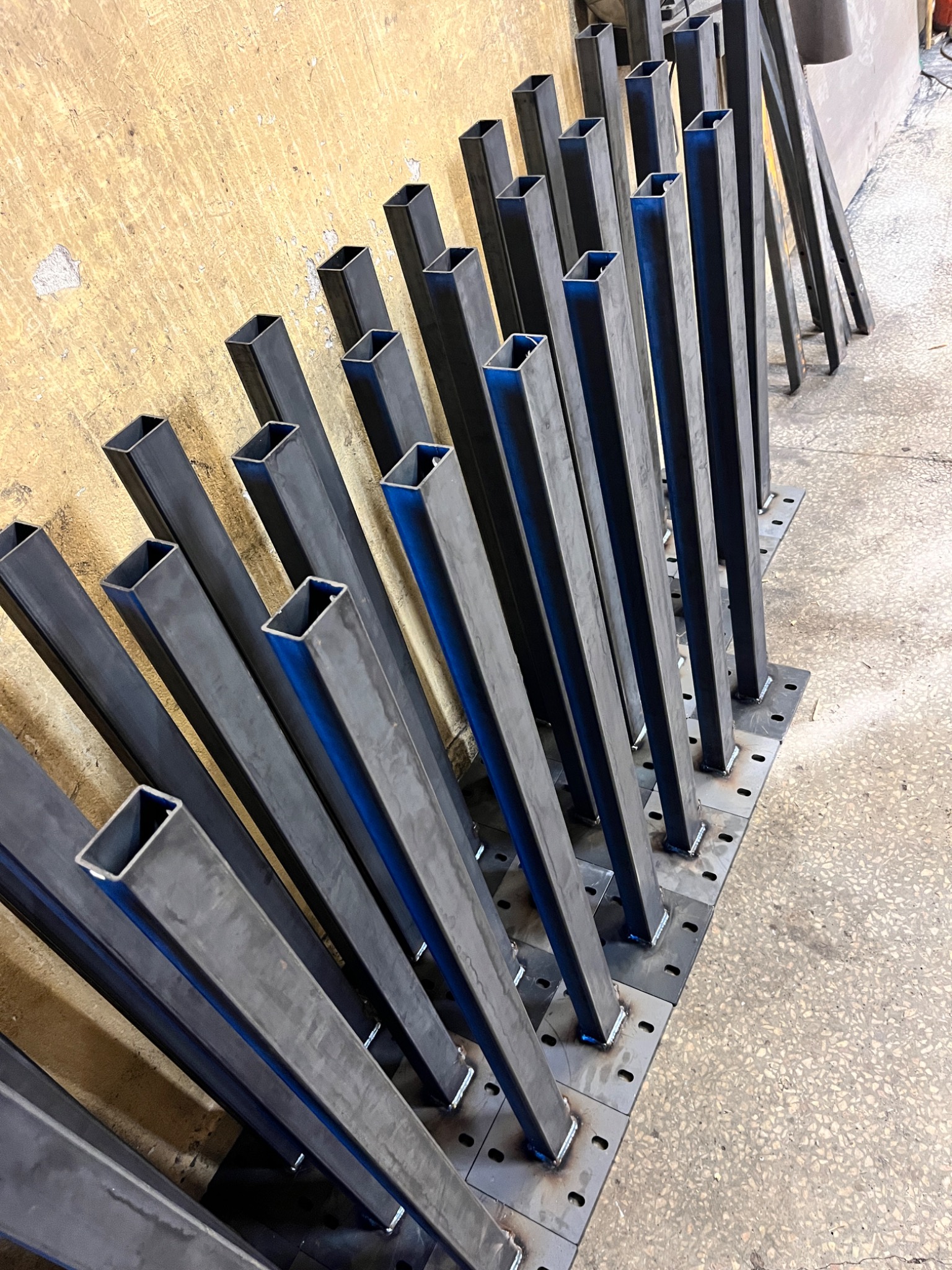 Przygotowane do montażu metalowe słupki o przekroju kwadratowym, przyspawane do kwadratowych podstaw z otworami, ustawione rzędem w warsztacie.