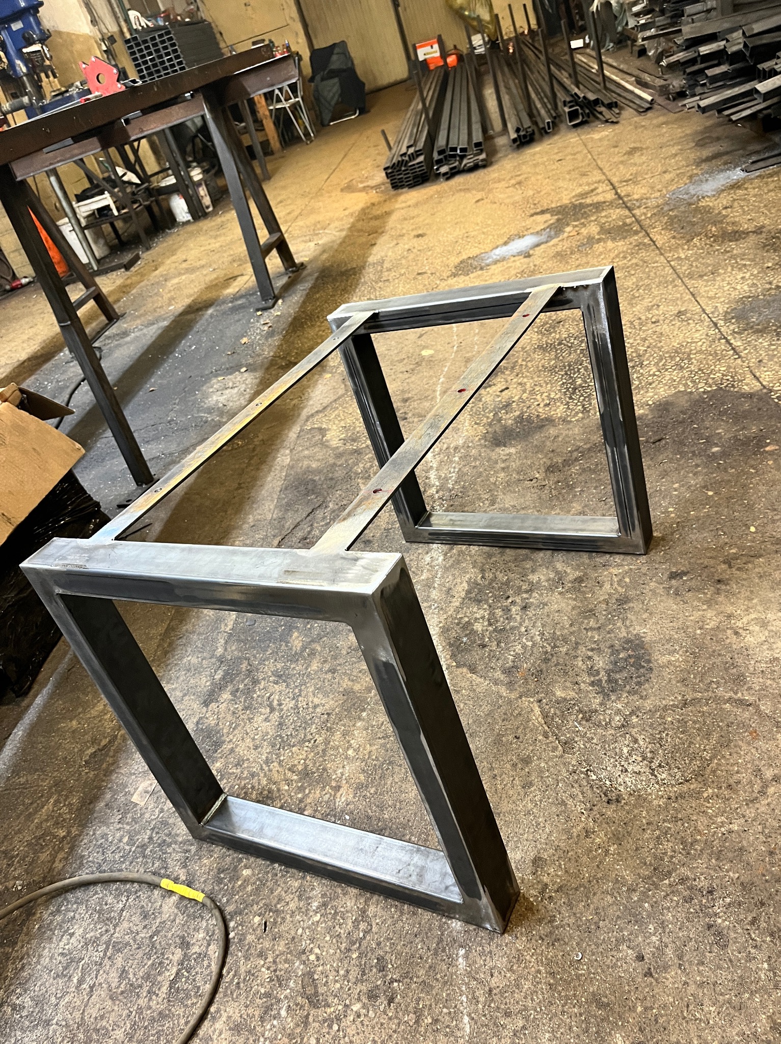 Dwie metalowe ramy w kształcie prostokątów połączone poprzeczką, leżące na betonowej podłodze w warsztacie. Widoczne ślady spawania i obróbki metalu, w tle profile stalowe i stanowisko pracy.