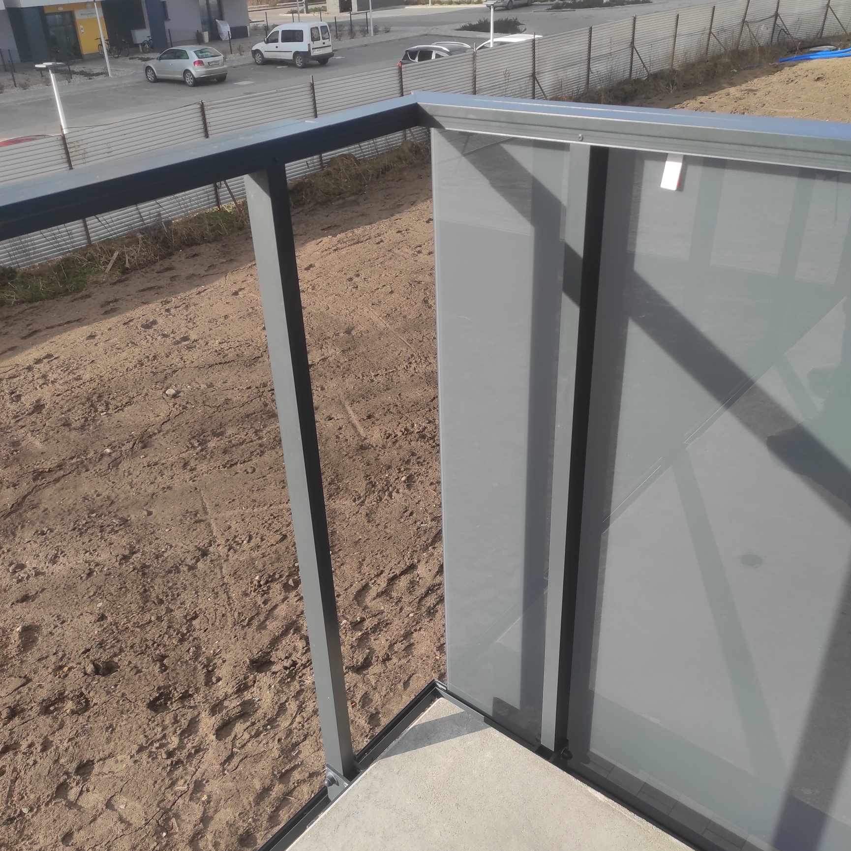 Szklana balustrada balkonowa z metalowymi, grafitowymi słupkami, widok z góry na plac budowy z zaparkowanymi samochodami w tle.