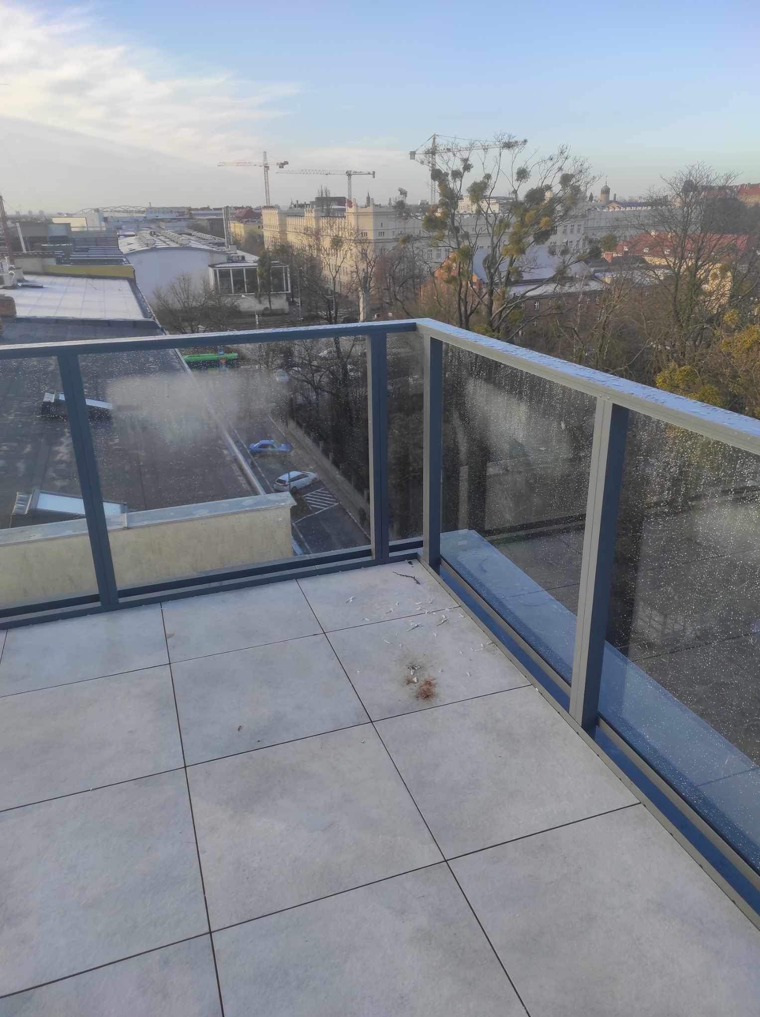 Szklana balustrada balkonowa z aluminiowymi ramami, widok na miasto w tle, lekko pochmurne niebo.