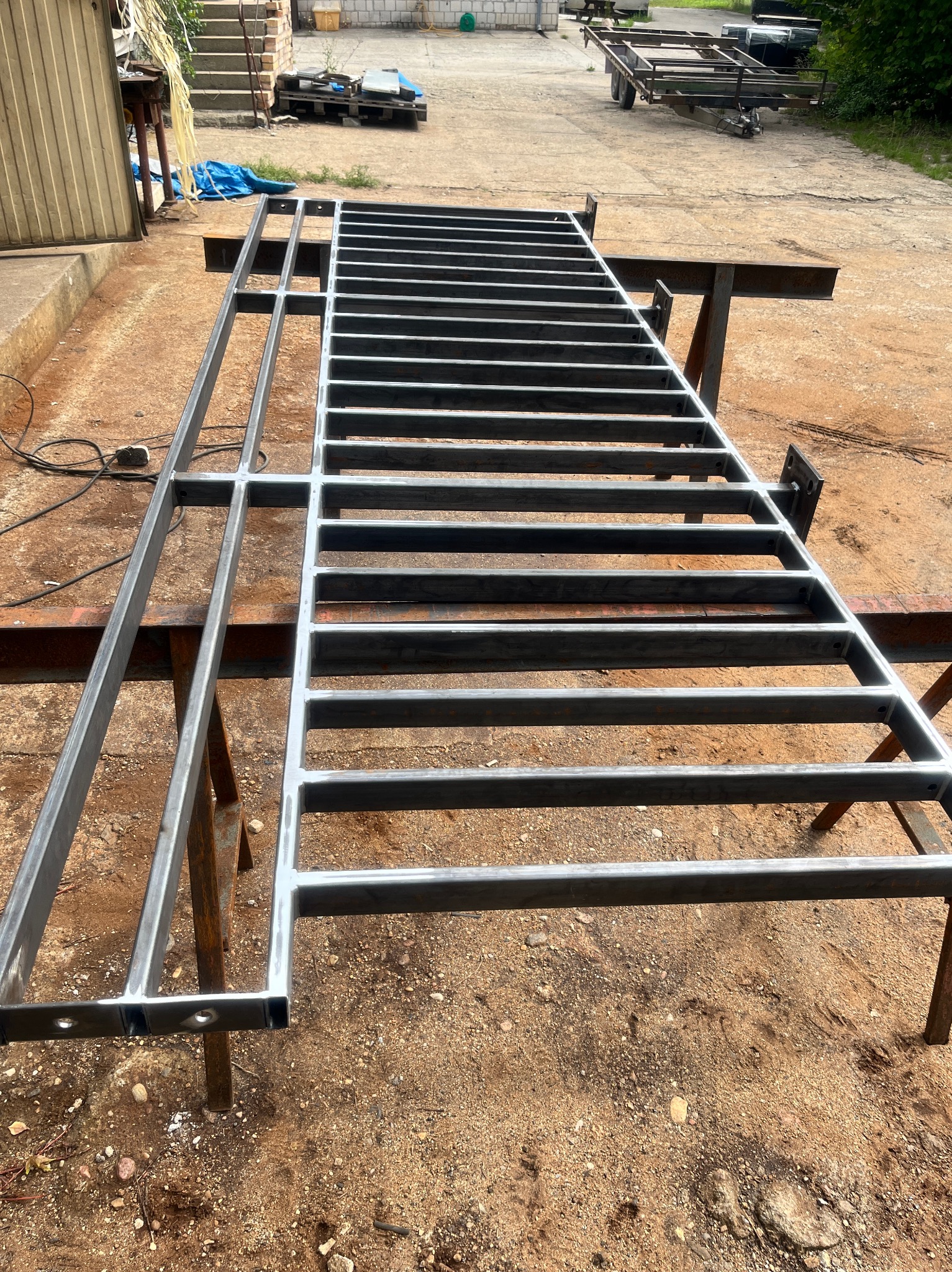 Surowa, metalowa konstrukcja kratowa z profili, wsparta na kozłach spawalniczych na zewnątrz, widoczne ślady spawania i przygotowania do dalszej obróbki.