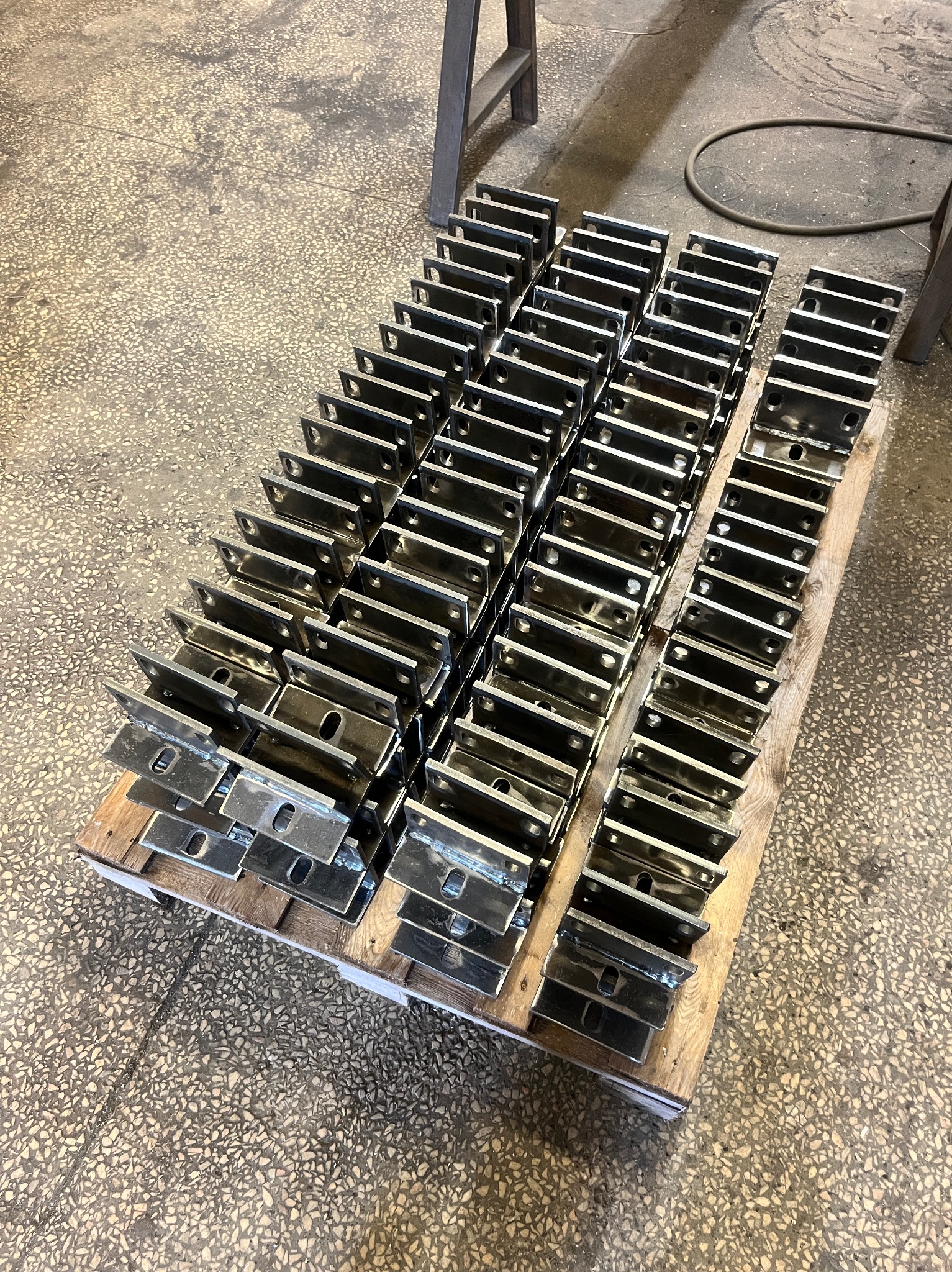 Partia metalowych uchwytów kątowych, ułożonych rzędami na drewnianej palecie w warsztatowym otoczeniu, widoczne ślady spawania.