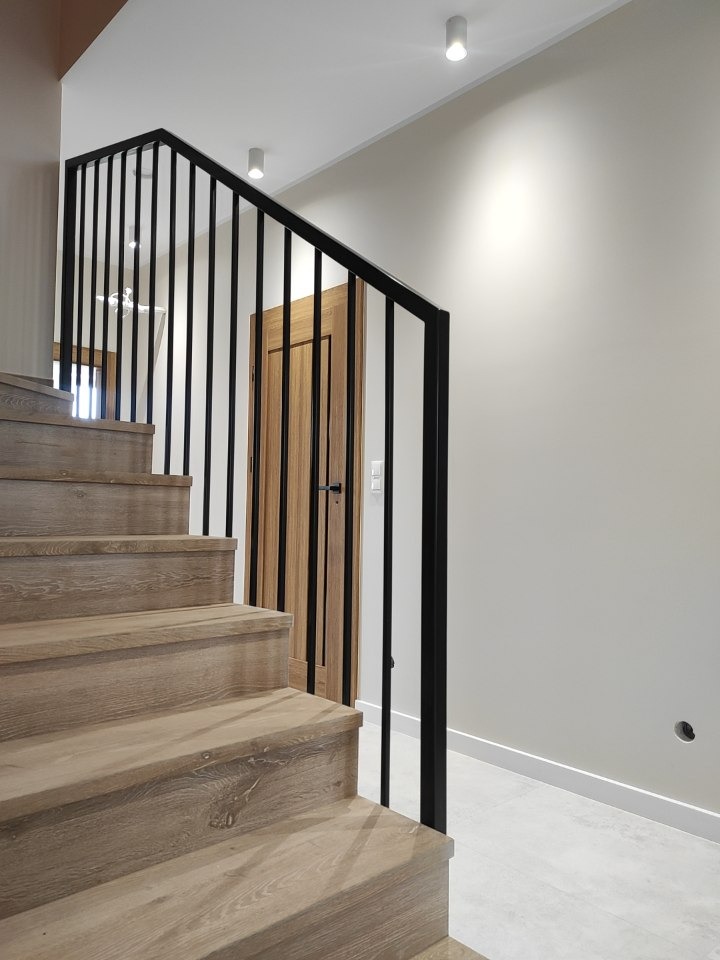 Drewniane schody z czarną, metalową balustradą w nowoczesnym wnętrzu, widok z dołu na kilka stopni i fragment drzwi w tle.
