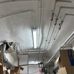 Aqua-Eco-Therm - Rozbudowana instalacja rur pod sufitem piwnicy z widocznym zbiornikiem i licznymi zaworami, oświetlona jarzeniówką.