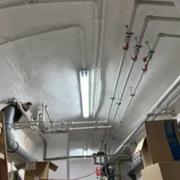 Rozbudowana instalacja rur pod sufitem piwnicy z widocznym zbiornikiem i licznymi zaworami, oświetlona jarzeniówką.