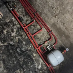 Czerwone rury instalacji grzewczej przymocowane do surowej, kamiennej ściany piwnicy, widoczny biały zbiornik wyrównawczy i manometr.