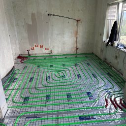 Aqua-Eco-Therm - Wnętrze w trakcie montażu ogrzewania podłogowego: zielone rurki ułożone na folii z siatką, widoczne przyłącza do instalacji na ścianie w surowym stanie.
