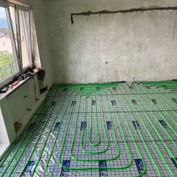 Aqua-Eco-Therm - Wnętrze w trakcie montażu ogrzewania podłogowego: widok na rozłożone, zielone rurki grzewcze na folii z nadrukiem, surowy stan ścian i okno z narzędziami.