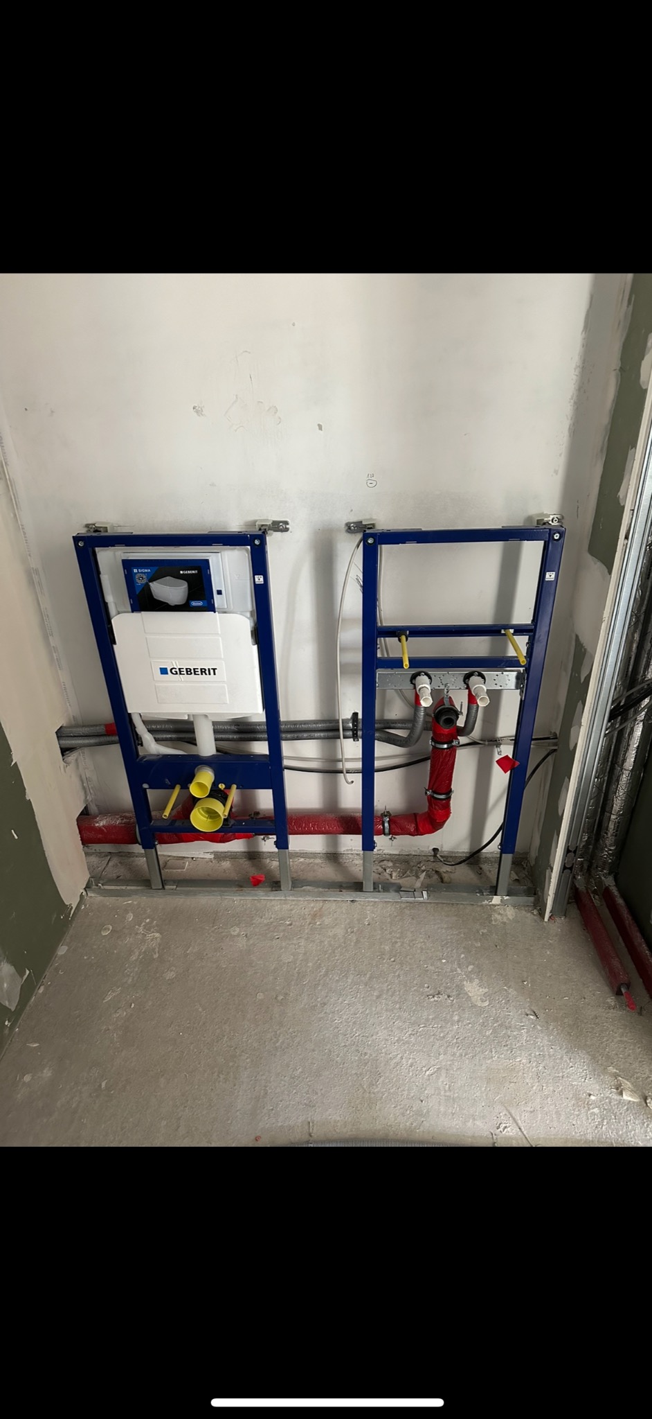Stelaż podtynkowy Geberit Duofix z częściowo podłączoną instalacją wodną w surowym stanie, widoczne rury w izolacji czerwonej.