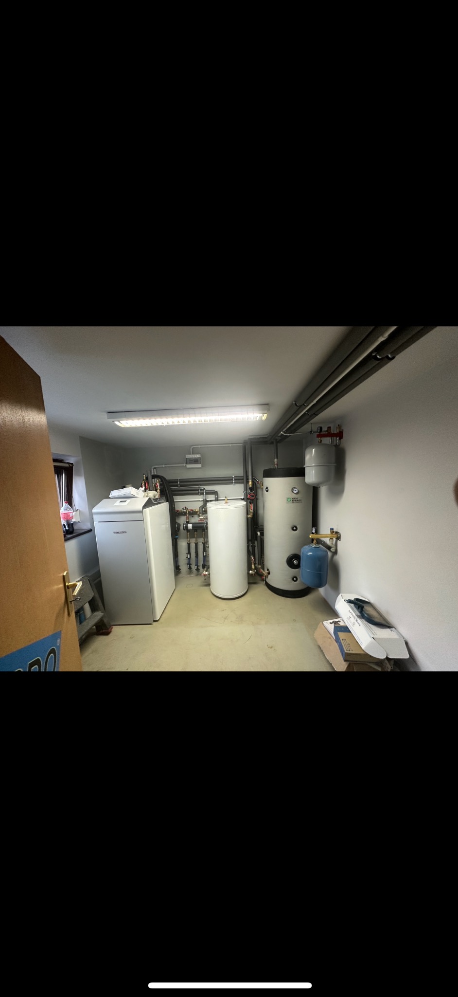 Kotłownia z dwoma zbiornikami akumulacyjnymi, nowoczesnym piecem i instalacją rurową pod sufitem, widoczne również kartony i butelka napoju.