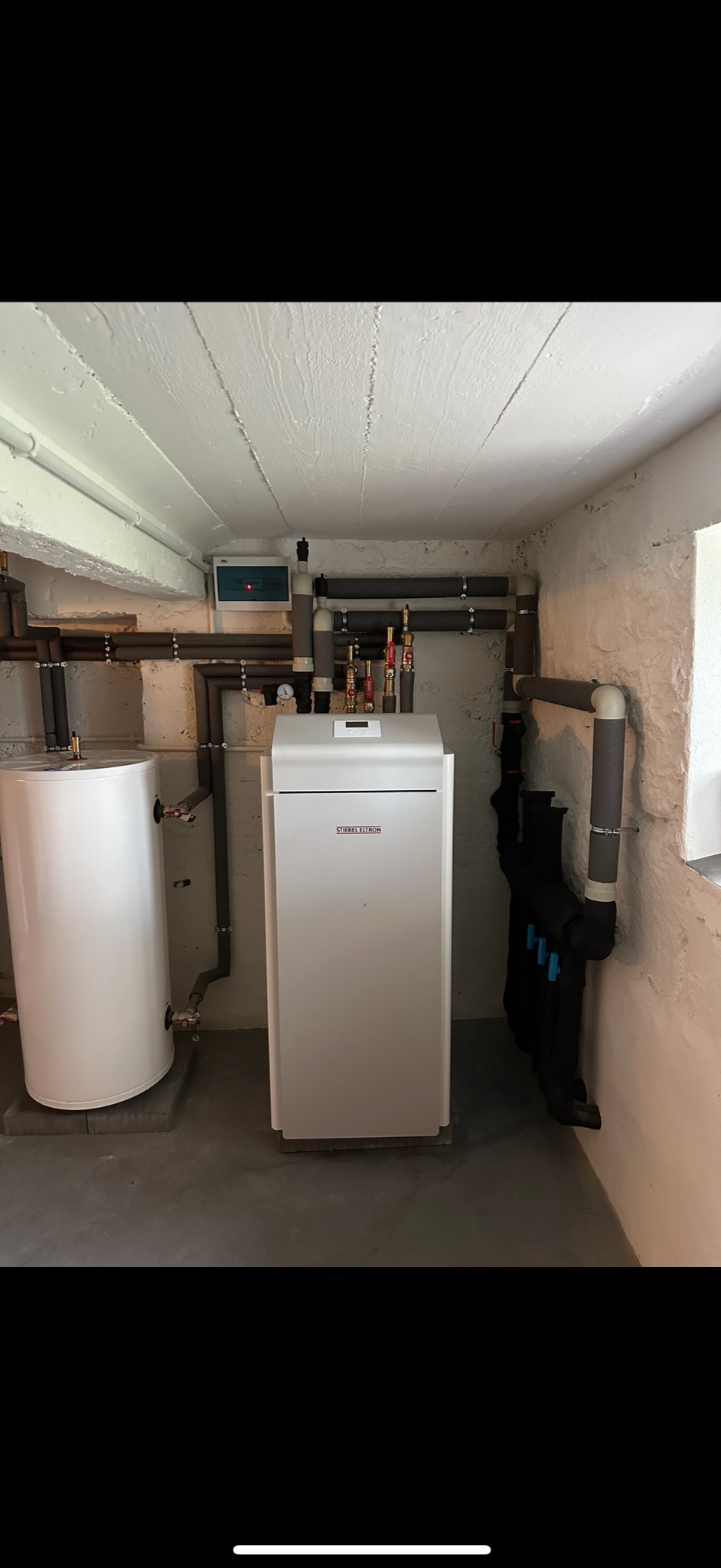 Kotłownia z białym zbiornikiem na wodę i piecem gazowym Stiebel Eltron, widoczne rury instalacji w piwnicy z surowymi ścianami.