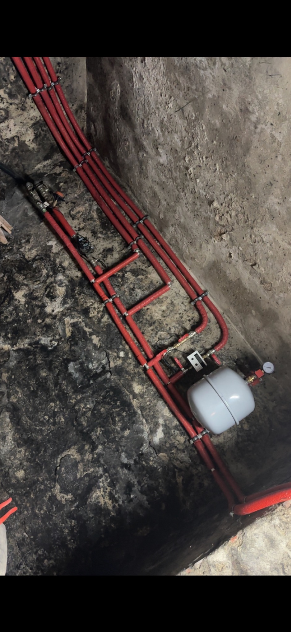 Czerwone rury instalacji grzewczej przymocowane do surowej, kamiennej ściany piwnicy, widoczny biały zbiornik wyrównawczy i manometr.