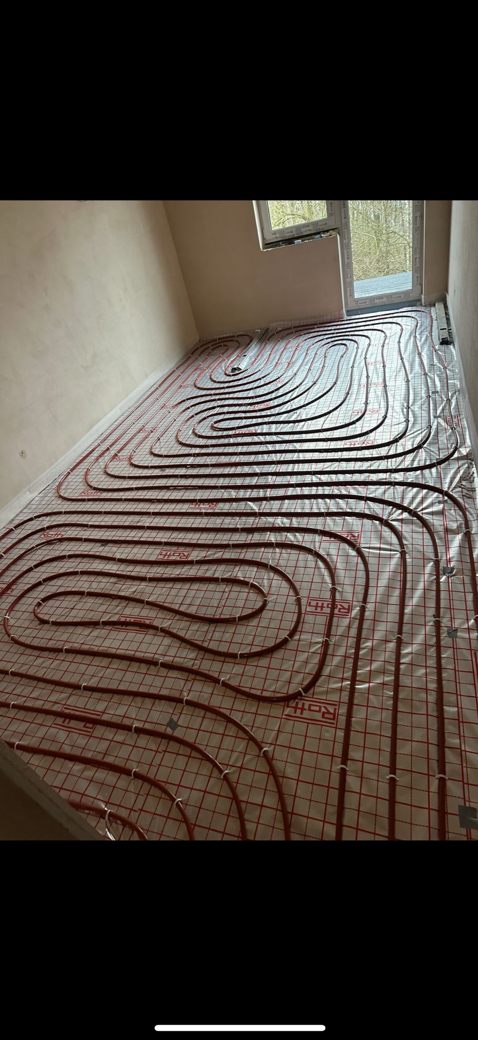 Rozłożona instalacja ogrzewania podłogowego Roth na folii z nadrukiem kratki, widoczne rury w kolorze ceglastym i białe uchwyty mocujące, surowy stan ścian i okno w tle.