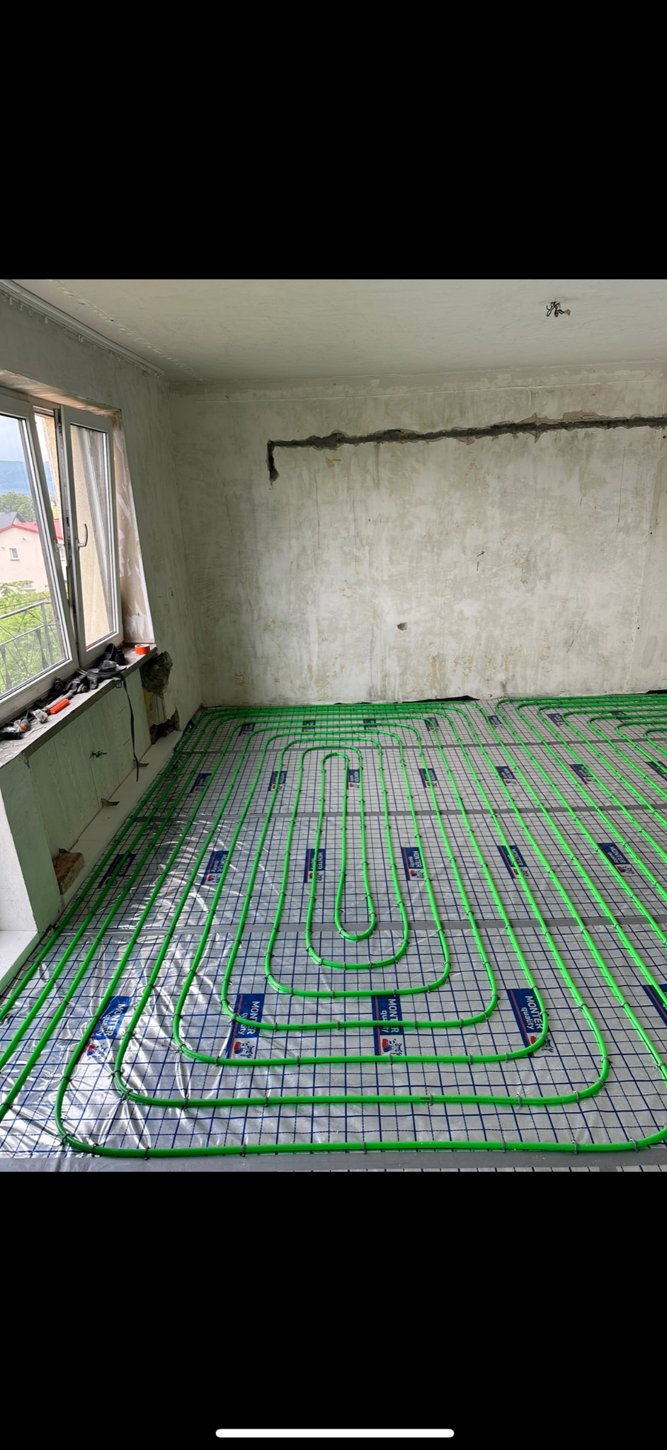 Wnętrze w trakcie montażu ogrzewania podłogowego: widok na rozłożone, zielone rurki grzewcze na folii z nadrukiem, surowy stan ścian i okno z narzędziami.
