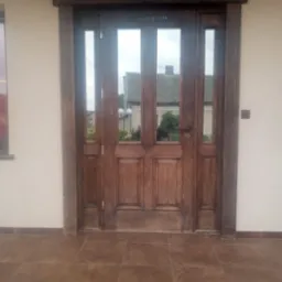 Odrestaurowane, drewniane drzwi wejściowe z szybami, ozdobnymi kolumnami po bokach i napisem 'K+B 2024' u góry, widoczne z zewnątrz na tle jasnej elewacji.