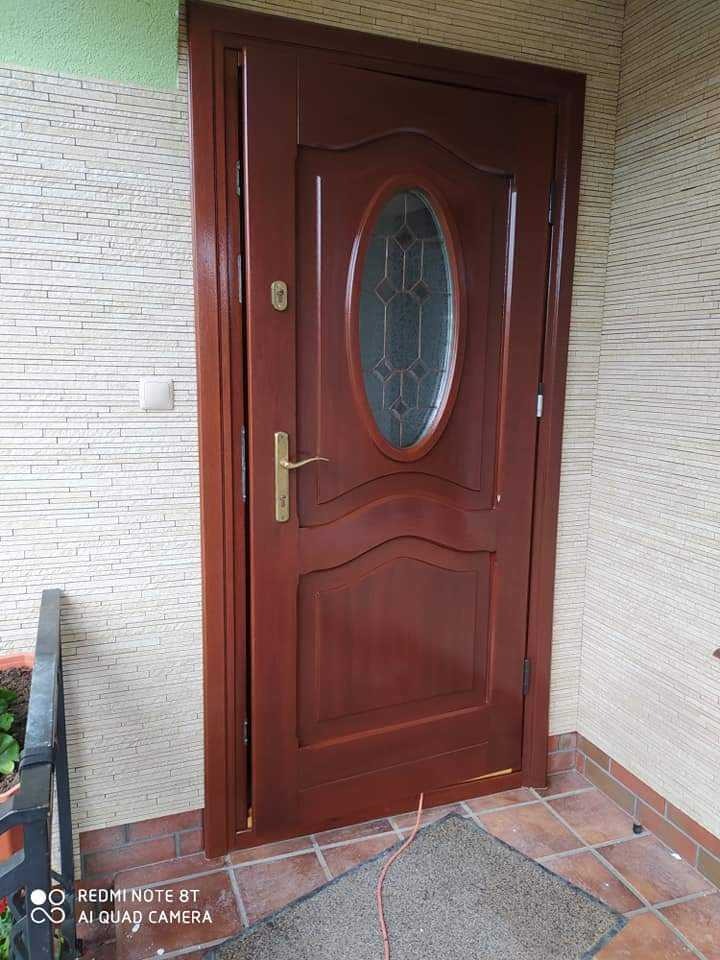 Ciemnobrązowe drewniane drzwi wejściowe z owalnym przeszkleniem, widoczne z zewnątrz, zamontowane w jasnej elewacji z dekoracyjną fakturą kamienia.