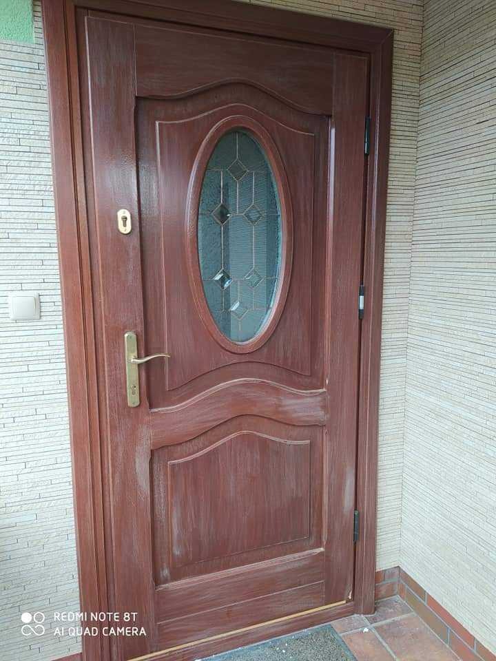 Brązowe drzwi wejściowe z owalnym przeszkleniem, po renowacji, widoczne ślady farby i niedociągnięcia w malowaniu.