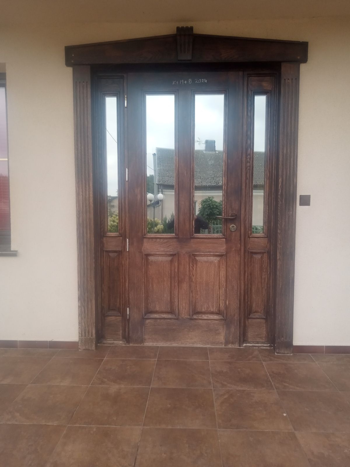 Odrestaurowane, drewniane drzwi wejściowe z szybami, ozdobnymi kolumnami po bokach i napisem 'K+B 2024' u góry, widoczne z zewnątrz na tle jasnej elewacji.