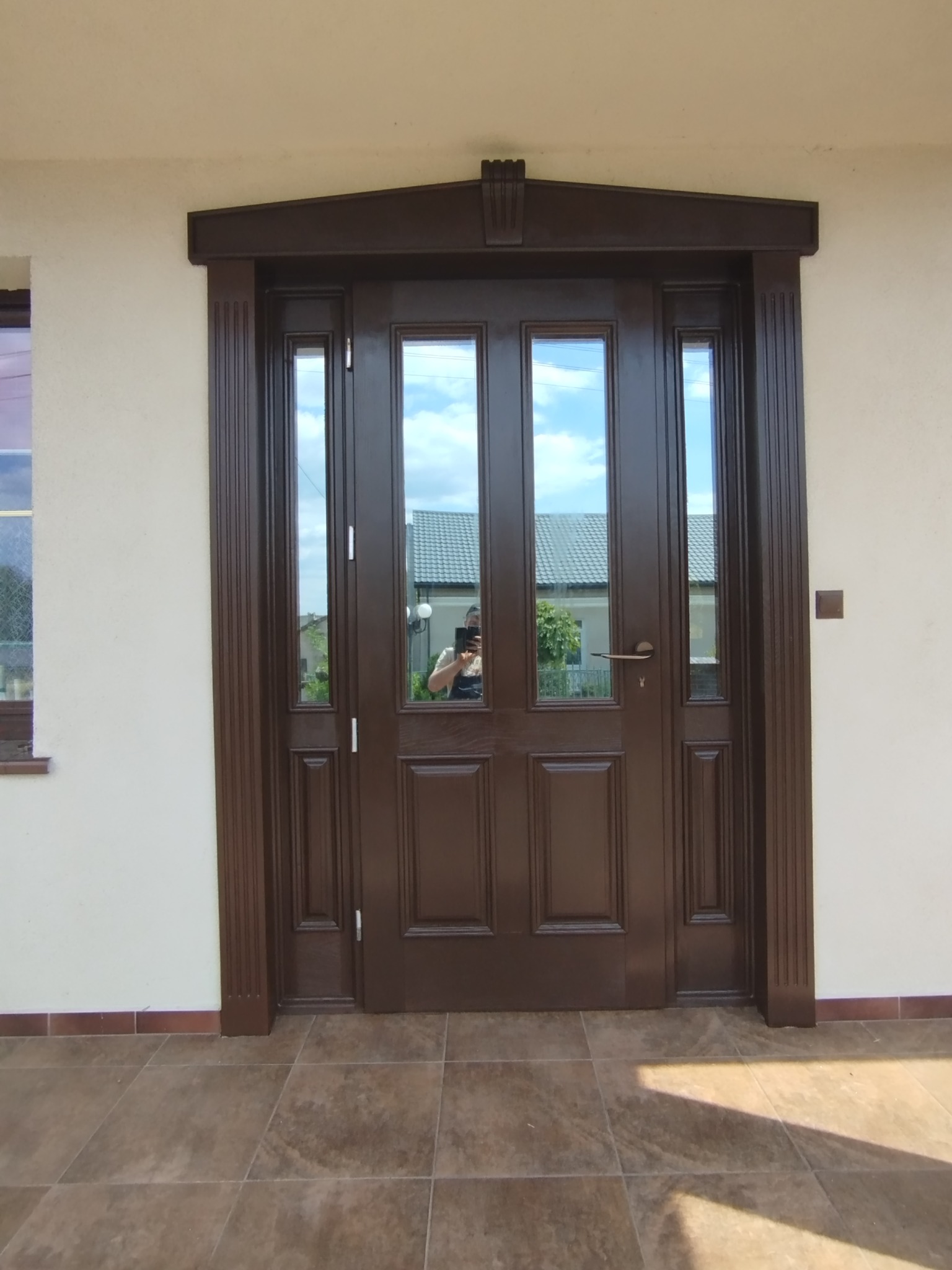 Eleganckie, brązowe drzwi wejściowe z szybami w Łomży, otoczone dekoracyjną ramą. W szybach odbija się budynek i niebo. Widoczne płytki na podłodze.