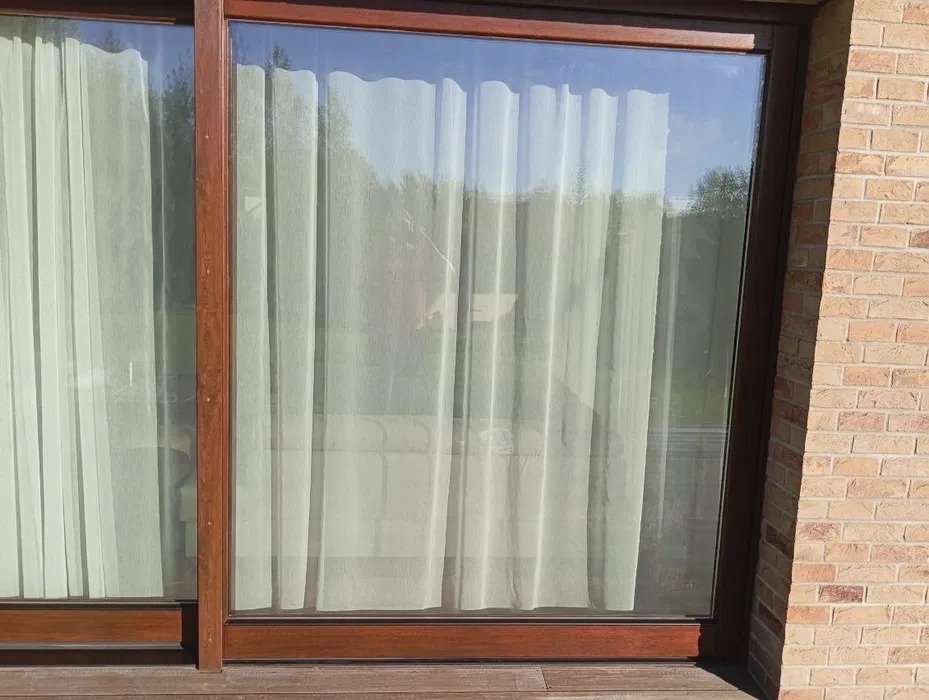 Przesuwne okno balkonowe z brązową ramą, widok na zewnątrz przez firankę. Część okna przylega do ściany z cegły. Widoczna drewniana podłoga balkonu.