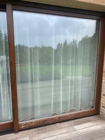 Drewniane okno przesuwne z widocznymi śladami zużycia na ramie, zasłonięte firanką, z widokiem na zieleń za oknem.