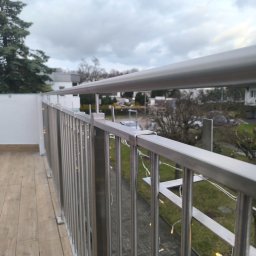 M-J-Stal Grzegorz Molenda - Balkon z podłogą z drewnopodobnych paneli i nowoczesną balustradą ze stali nierdzewnej, ozdobioną delikatnymi lampkami sznurkowymi, widok na osiedle w pochmurny dzień.