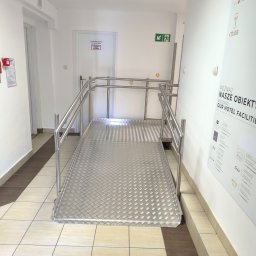 M-J-Stal Grzegorz Molenda - Metalowa rampa dla wózków inwalidzkich w hotelowym korytarzu, z poręczami i antypoślizgową powierzchnią. Widoczny plakat reklamowy hotelu na ścianie.