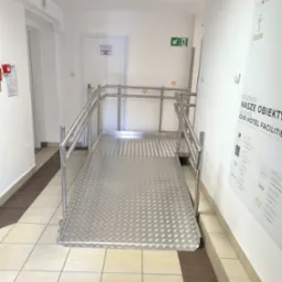 Metalowa rampa dla wózków inwalidzkich w hotelowym korytarzu, z poręczami i antypoślizgową powierzchnią. Widoczny plakat reklamowy hotelu na ścianie.
