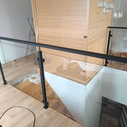 M-J-Stal Grzegorz Molenda - Szklana balustrada z czarnymi elementami mocującymi zabezpiecza schody. Widoczne drewniane wykończenie podłogi i schodów. W tle drzwi i drabina.