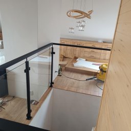 M-J-Stal Grzegorz Molenda - Nowoczesna balustrada szklana z czarnymi słupkami, widok z góry na jasne wnętrze z drewnianymi elementami i ozdobnym oświetleniem. Nowoczesny design.