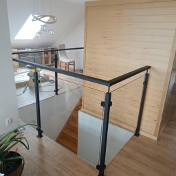 M-J-Stal Grzegorz Molenda - Szklana balustrada z czarnymi, metalowymi słupkami w nowoczesnym wnętrzu. Widoczny fragment schodów, drewniana boazeria i roślina doniczkowa.