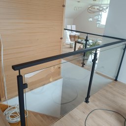 M-J-Stal Grzegorz Molenda - Nowoczesna balustrada szklana z czarnymi elementami metalowymi, zamontowana na schodach wewnątrz domu. Widoczne drewniane wykończenia i jasne wnętrze.