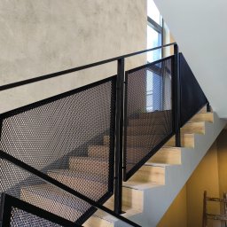 M-J-Stal Grzegorz Molenda - Nowoczesne schody z drewnianymi stopniami i czarną, metalową balustradą z siatki, widziane z dołu, z fragmentem okna w tle. Minimalistyczny design.