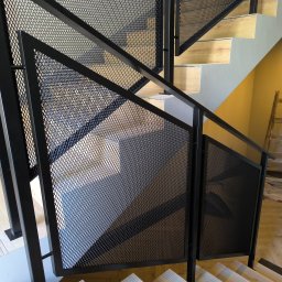 M-J-Stal Grzegorz Molenda - Nowoczesne schody z czarną, metalową balustradą z siatki o romboidalnych oczkach. Jasne, drewniane stopnie. Ujęcie z dołu, podkreślające geometrię konstrukcji.