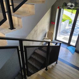 M-J-Stal Grzegorz Molenda - Nowoczesne schody z drewnianymi stopniami i czarną, metalową balustradą z siatki, widziane z góry. W tle jasne wnętrze z krzesłem i wyjściem na zewnątrz.