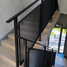 M-J-Stal Grzegorz Molenda - Schody z drewnianymi stopniami i czarną, metalową balustradą z siatkowym wypełnieniem. Widok na drzwi wejściowe i zewnętrzny chodnik. Nowoczesny design.