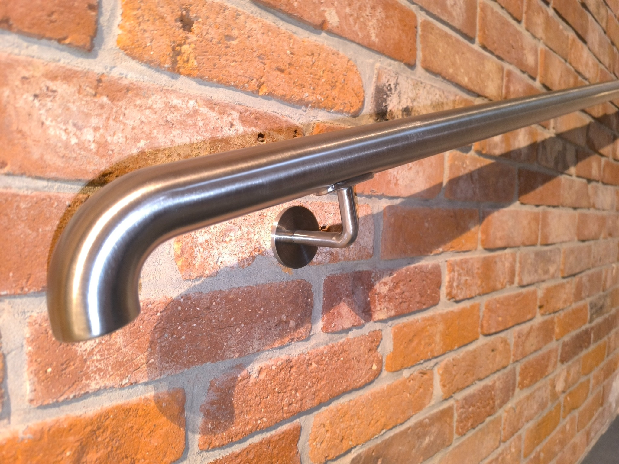 Nierdzewna poręcz przyścienna, zamocowana na ceglanej ścianie, z widocznymi detalami mocowania. Poręcz o satynowym wykończeniu.