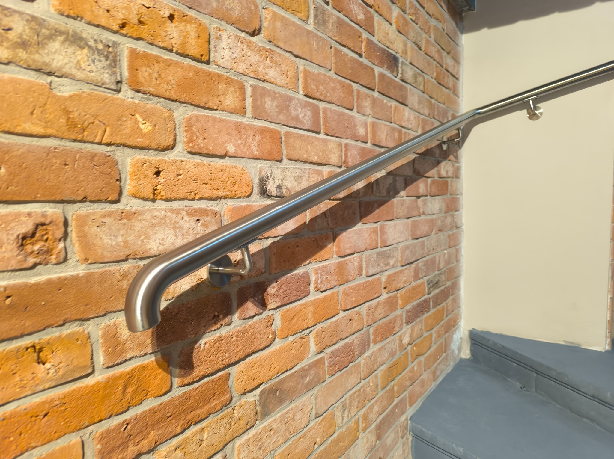 Metalowa poręcz schodowa o gładkim wykończeniu, zamocowana na ceglanej ścianie. Widok z dołu, podkreślający solidność konstrukcji i estetyczne połączenie materiałów.