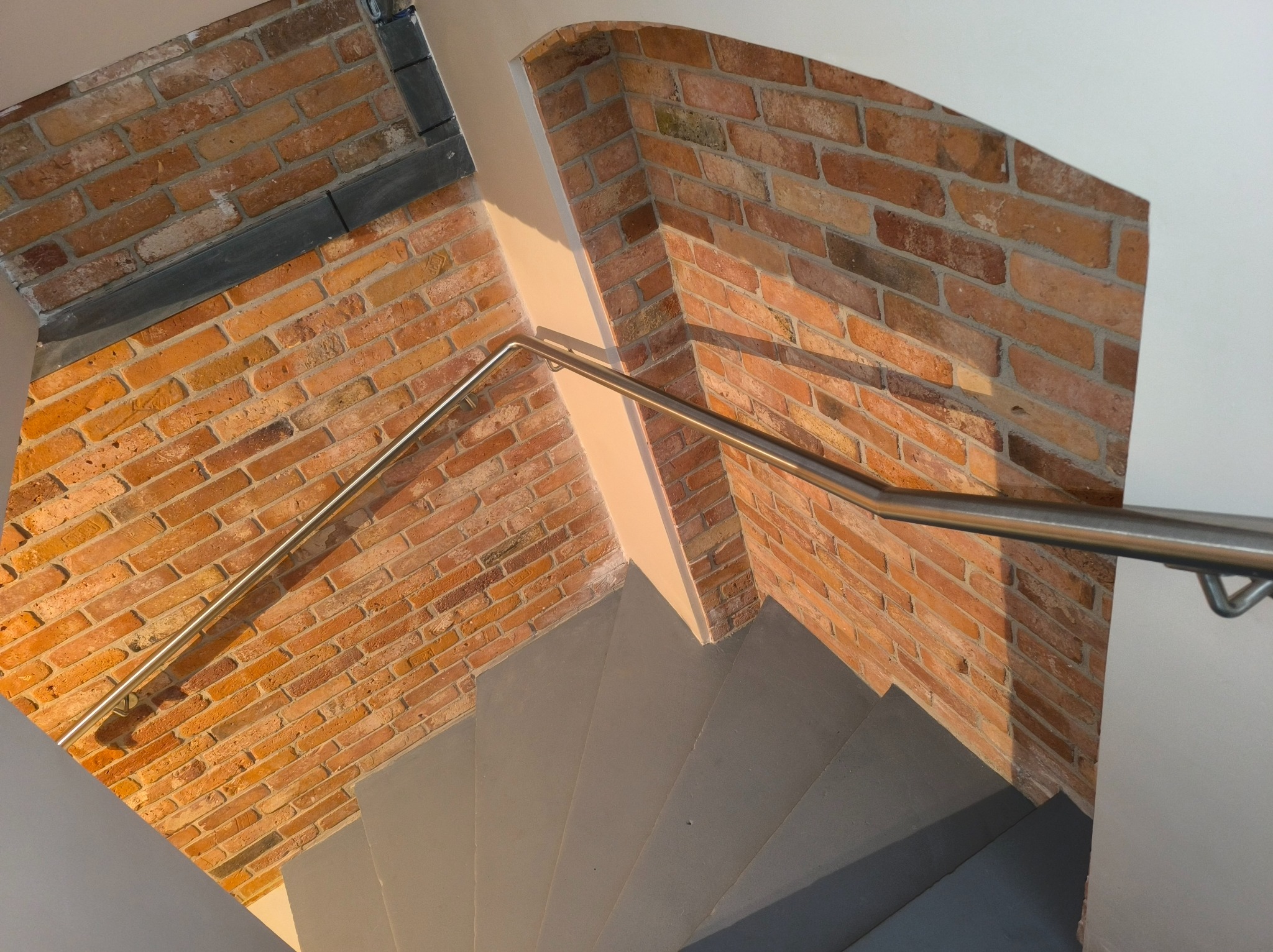Wewnętrzne schody z szarymi stopniami i metalową poręczą, obok ściana z cegły. Ujęcie z góry, widoczne fragmenty schodów i ściany.