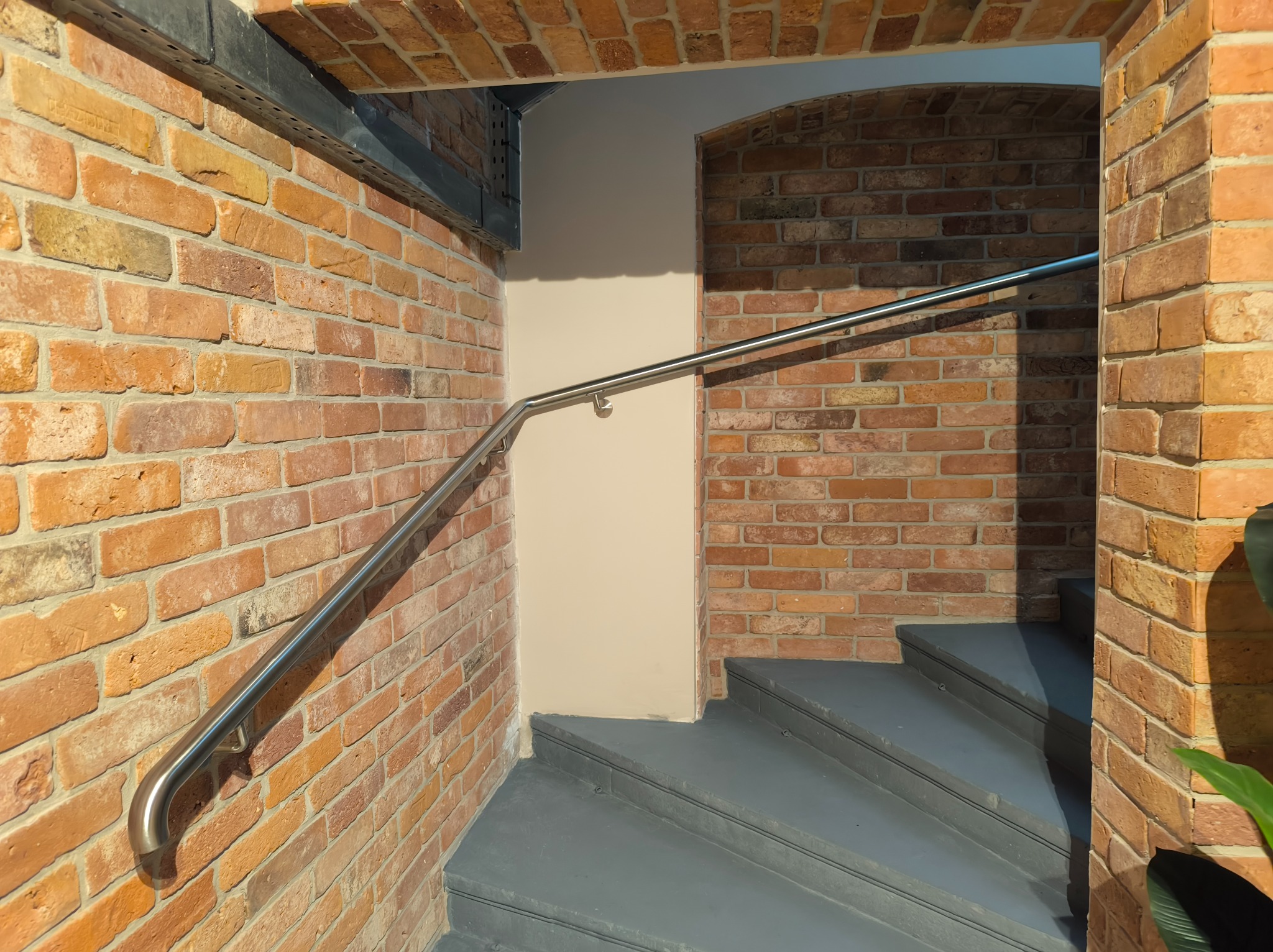 Wnętrze z ceglanymi ścianami i szarą klatką schodową. Metalowa poręcz zamontowana na ścianie prowadzi wzdłuż schodów. Charakterystyczna ceglana architektura.
