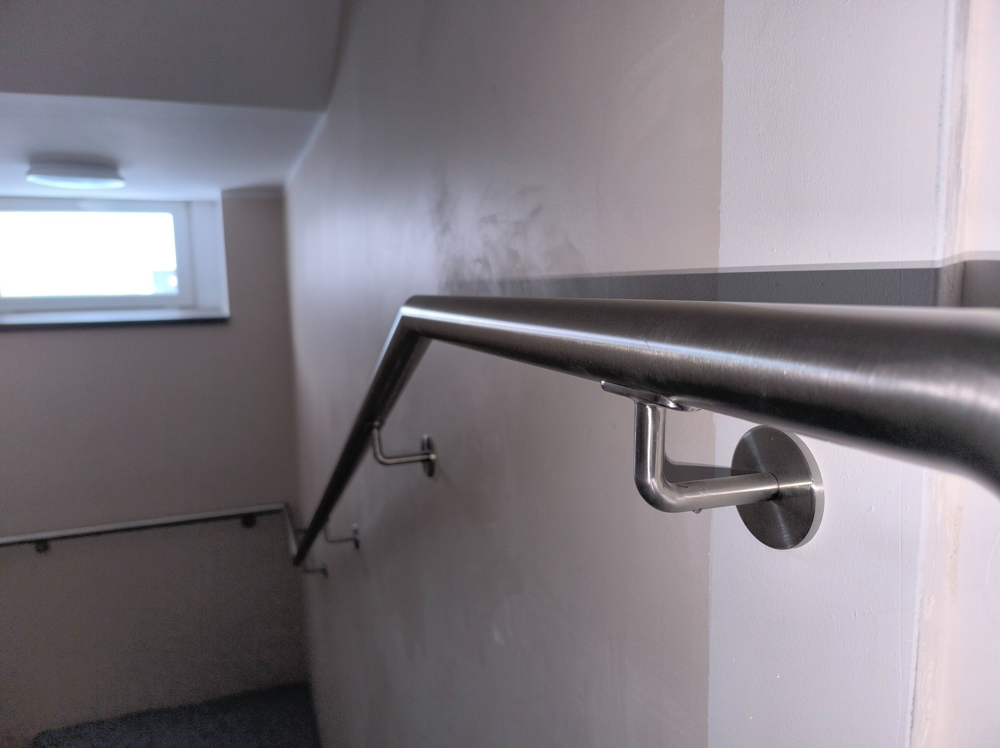 Metalowa poręcz przy ścianie w korytarzu, zamocowana na okrągłych uchwytach. Widoczne schody w tle i okno z białymi ramami, oświetlone światłem dziennym.