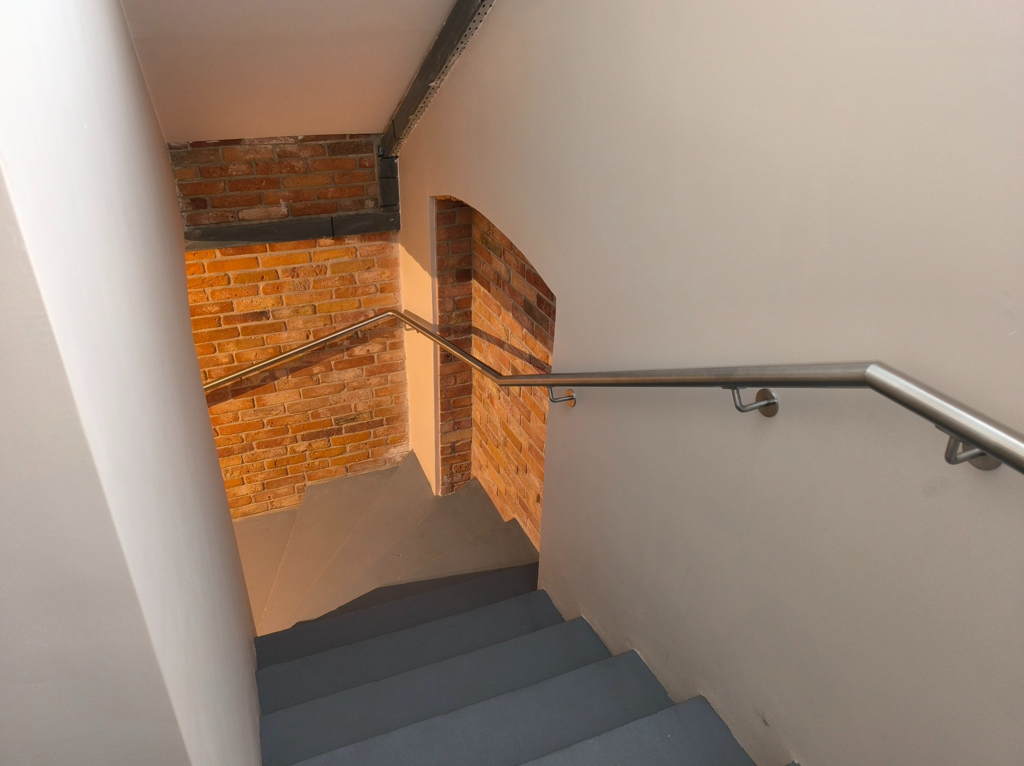 Wnętrze klatki schodowej z szarymi schodami i metalową poręczą, widok z góry. Ściany białe, fragment ściany z cegły. Oświetlenie naturalne.